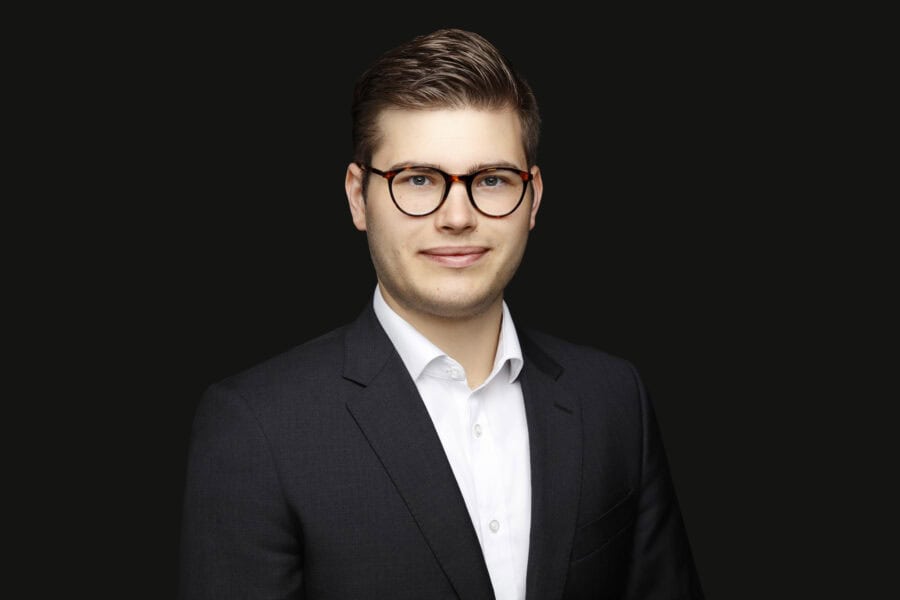 A photograph of Alex Jost wearing glasses and a black suit with a white shirt smiling slightly.