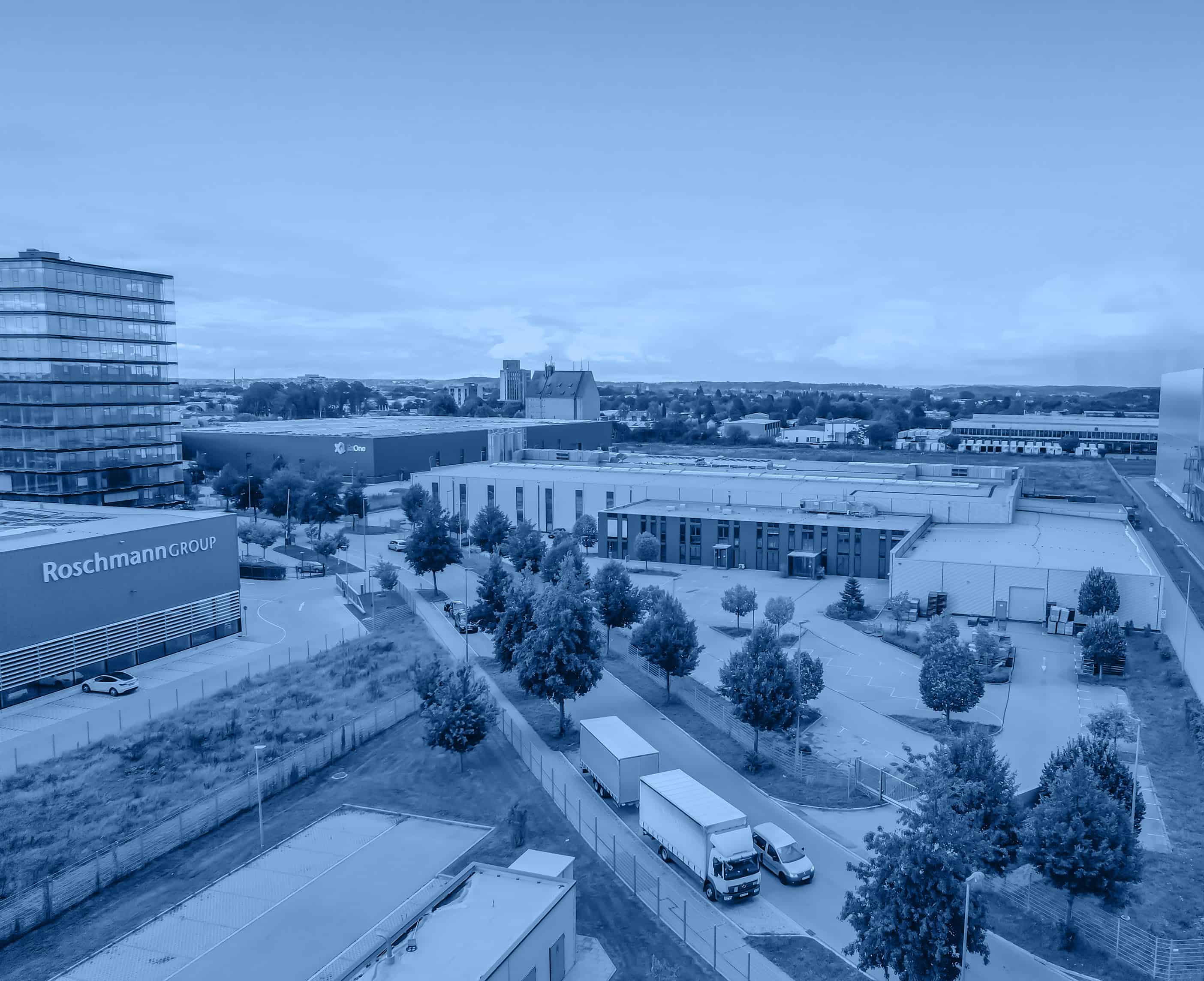 Industrial park featuring a large warehouses and a multi-story office building. Trucks on a road indicate active logistics. Trees line pathways, adding greenery.