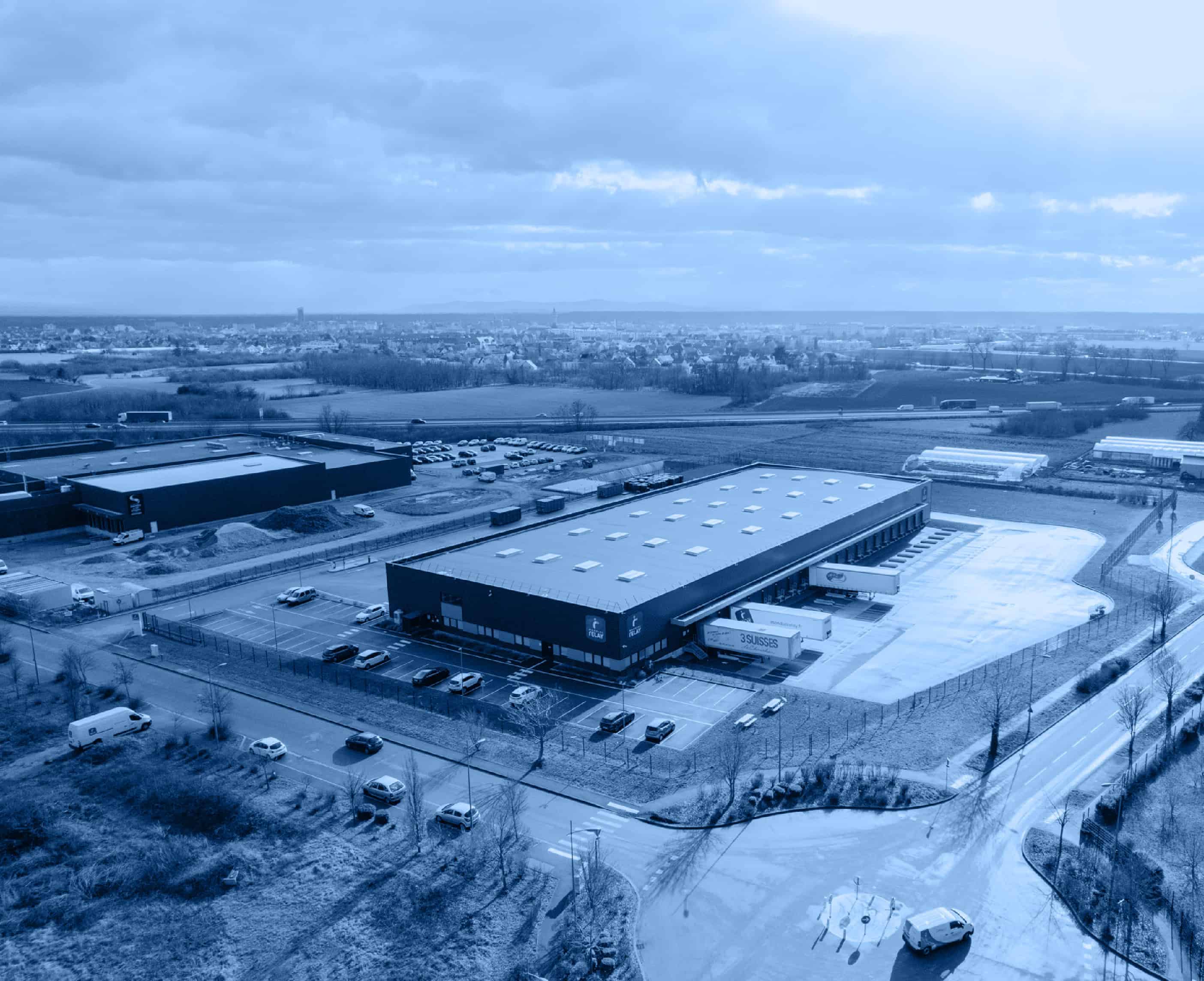 A large industrial warehouse with trucks parked outside on a cloudy day. Surrounding landscape features open fields and distant buildings.