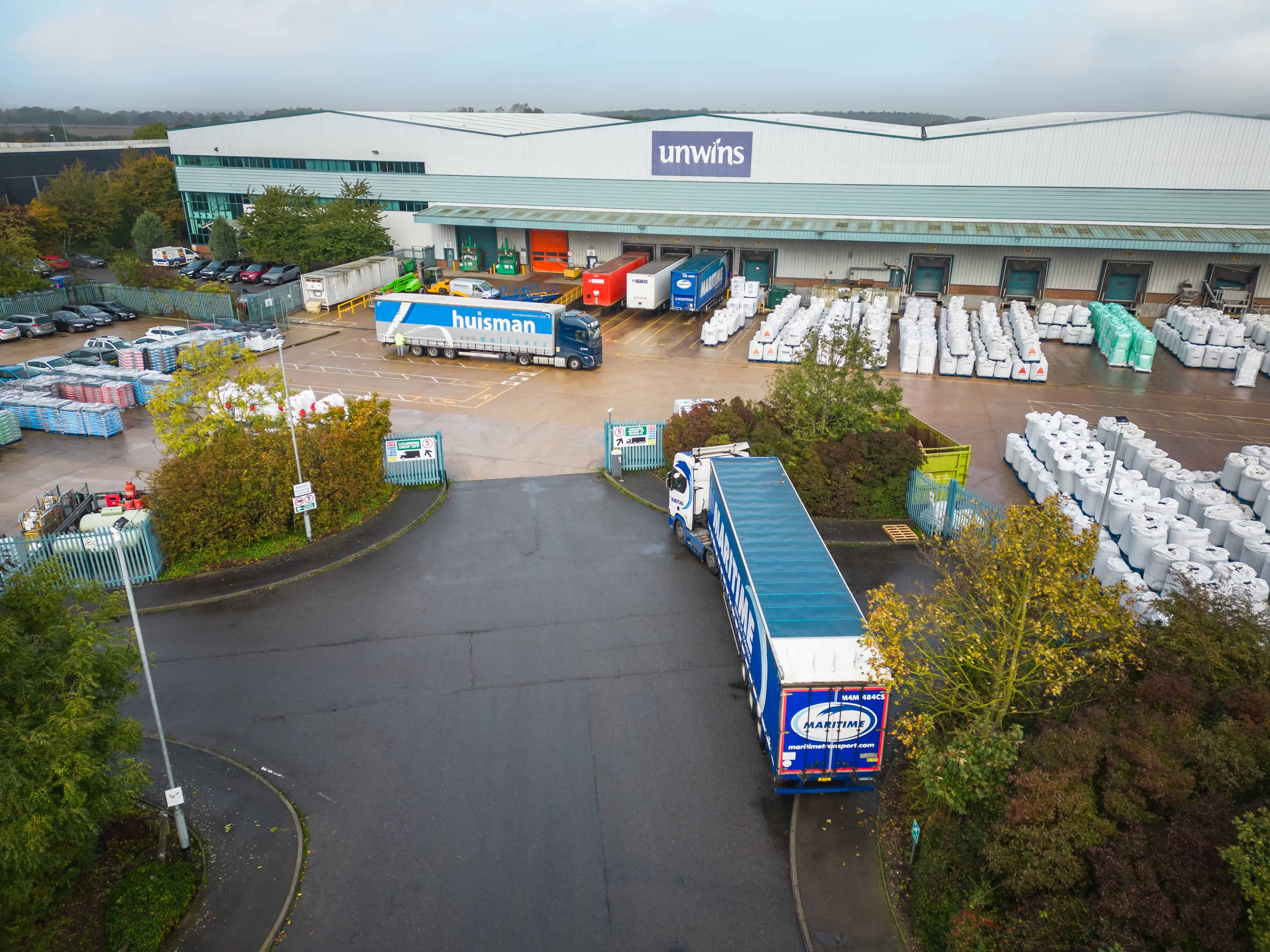 Aerial view of a large warehouse with the "Unwins" sign, surrounded by trucks and stacked goods. The scene is industrial, busy, and overcast.