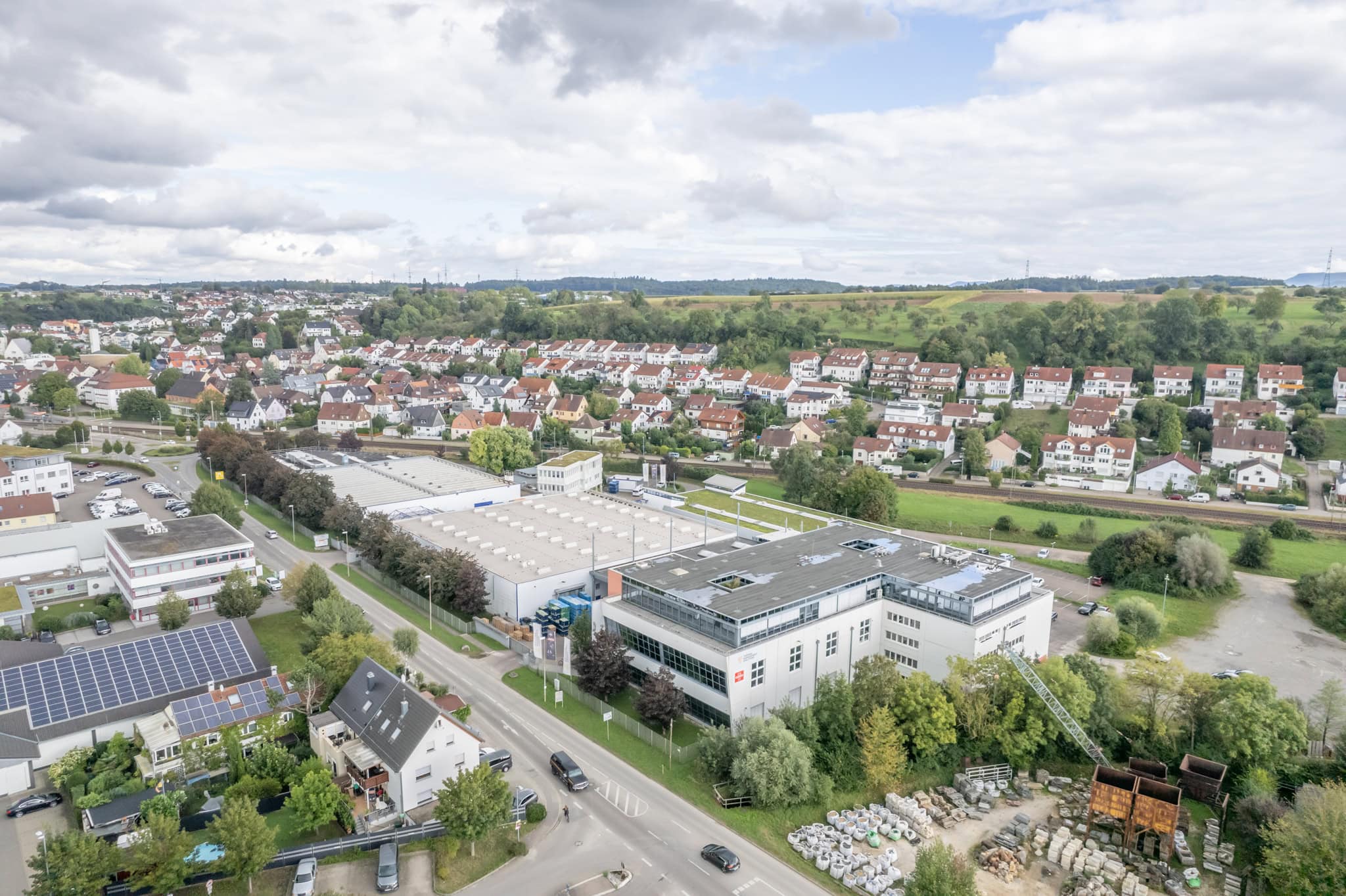 Aerial view of a suburban area with a mix of residential houses and industrial buildings, surrounded by greenery under a partly cloudy sky.