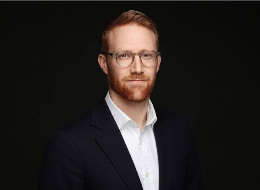 A photo of Peter Day wearing glasses and a dark suit against a black background.
